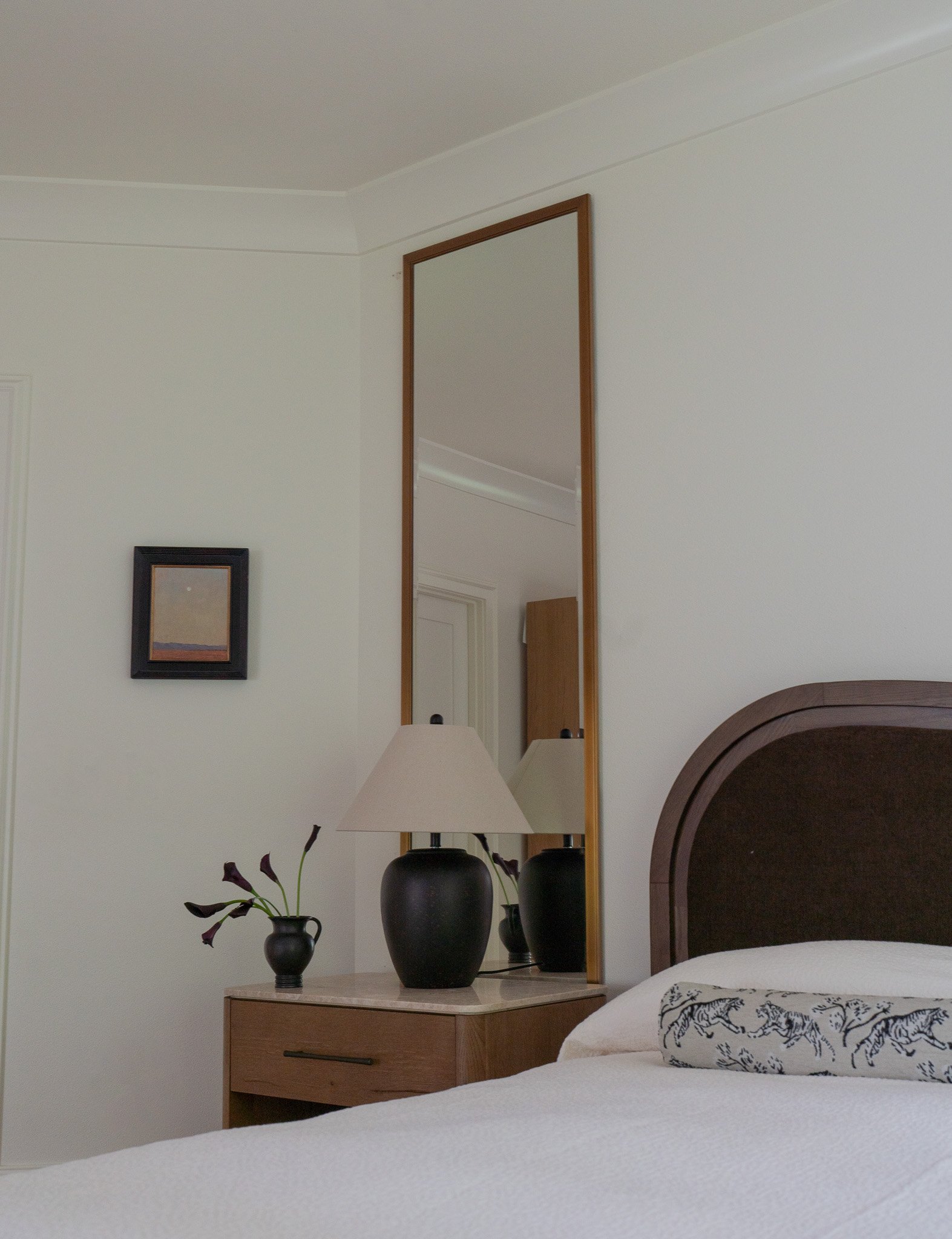 A bedroom with a white bed, a wooden nightstand holding three vases with dark purple flowers, a tall mirror on the wall, a framed picture, a table lamp, and part of a headboard.