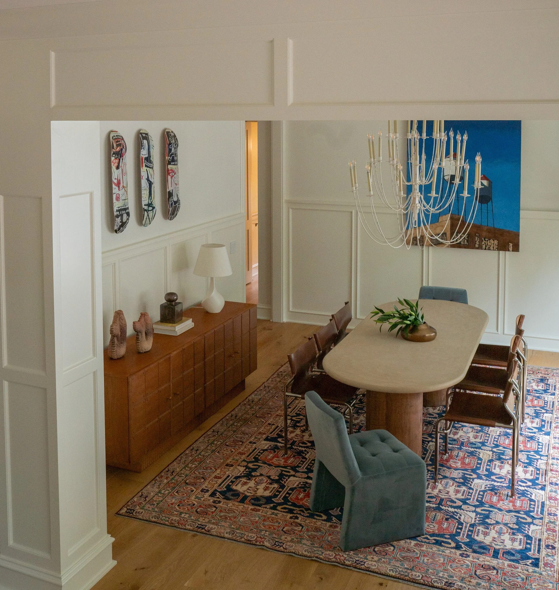 A dining room with a large ornate rug, a wooden sideboard, a round table with greenery in a vase, and blue chairs. On the wall, three skateboard decks are mounted, and a chandelier hangs from the ceiling. Part of a brightly painted room with artwork 