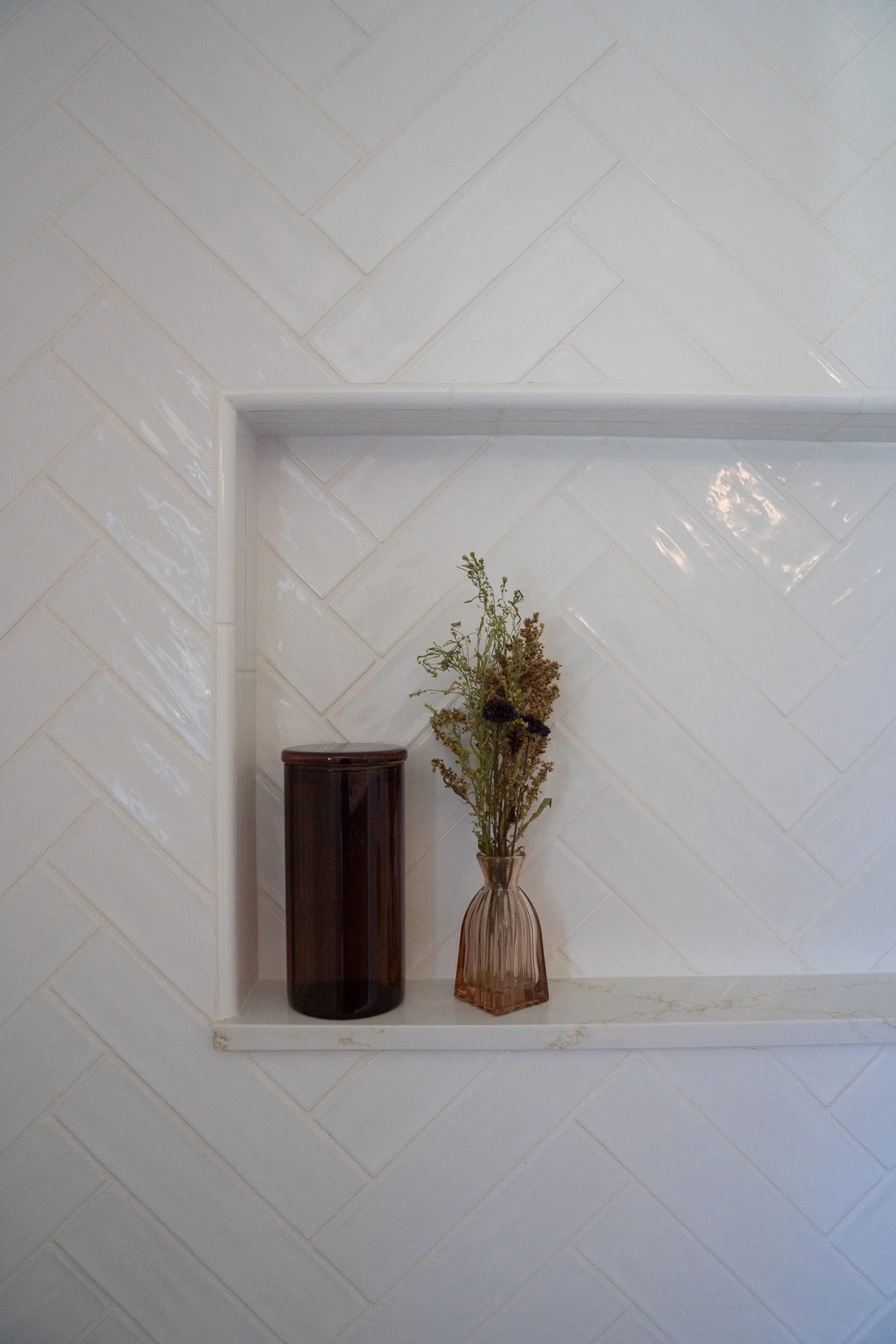 A white tiled wall with a built-in wall shelf holding a tall, dark brown glass vase and a shorter, amber-colored glass vase with dried flowers.