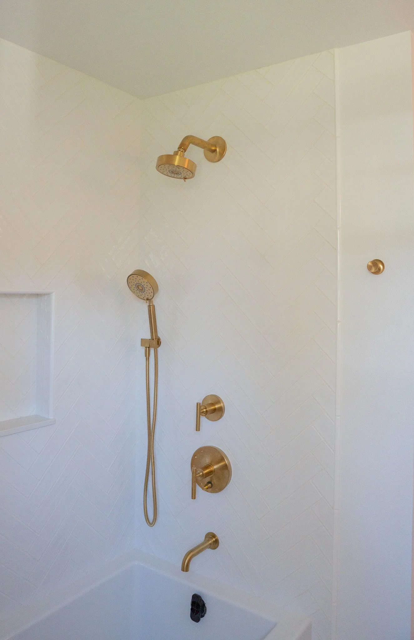 A white shower wall with gold shower fixtures, including a showerhead, hand shower, faucet, and a set of three controls, in a modern bathroom.