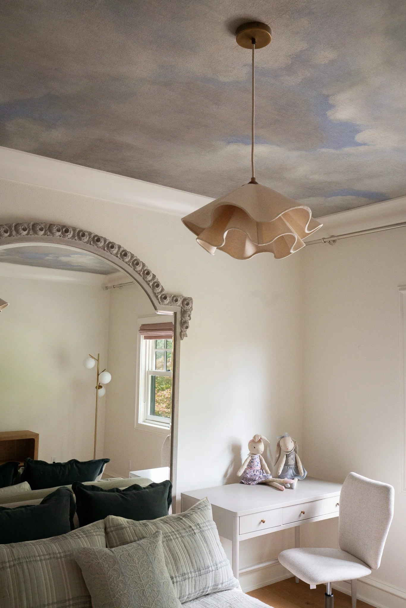 Living room with a white dresser on the wall and two plush bunny dolls on top, a white chair, a cream-colored sofa with multiple pillows, a window with blinds, a decorative mirror, and a ceiling light fixture with a wavy lampshade.