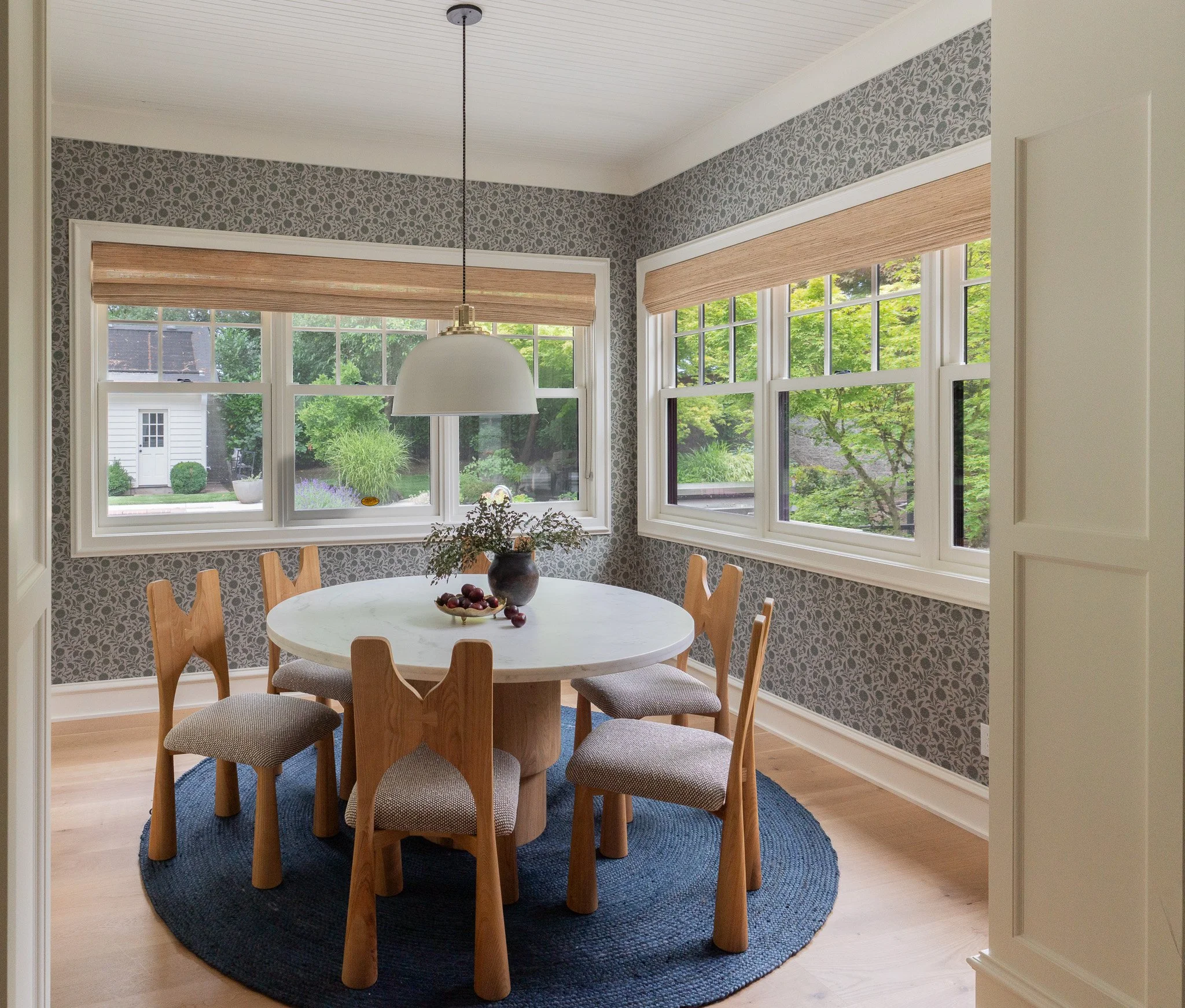 A bright dining area with large windows overlooking greenery. The room features a round white table with wooden chairs, a dark blue rug, and patterned wallpaper. A white pendant light hangs over the table.