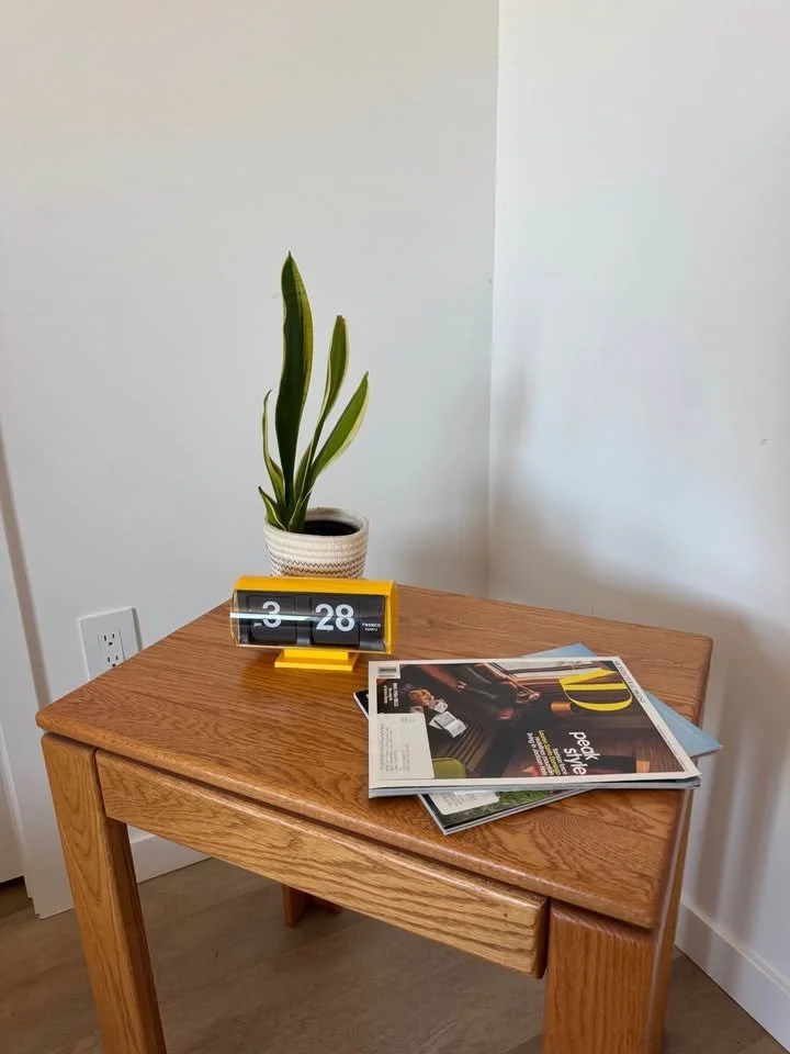 A wooden side table with a potted snake plant, a yellow flip clock displaying 3:28, and magazines on top, situated against a white wall with an electrical outlet.