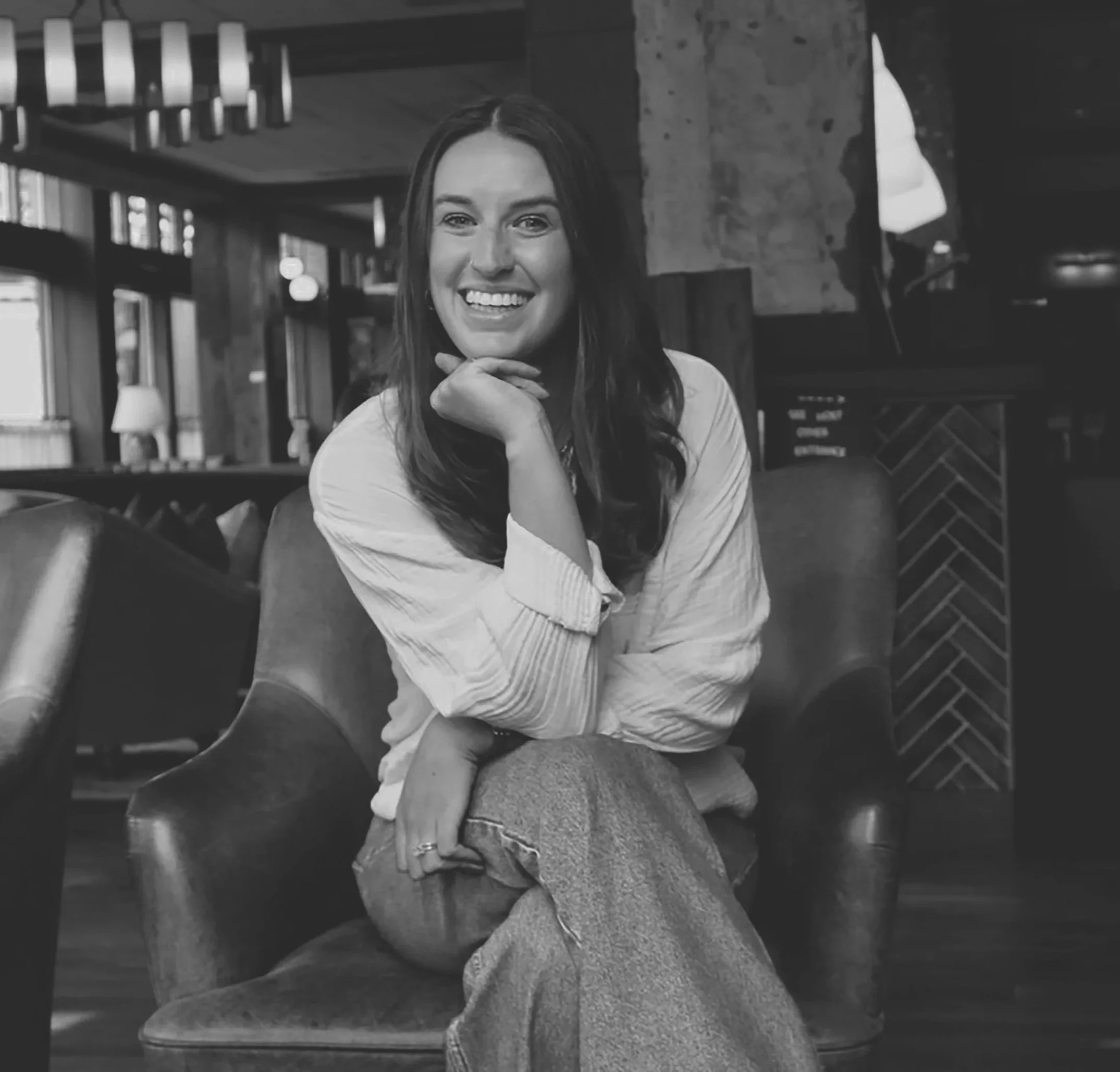 A woman with long dark hair smiles and rests her chin on her hand, sitting indoors in front of a bookshelf and a brick wall.