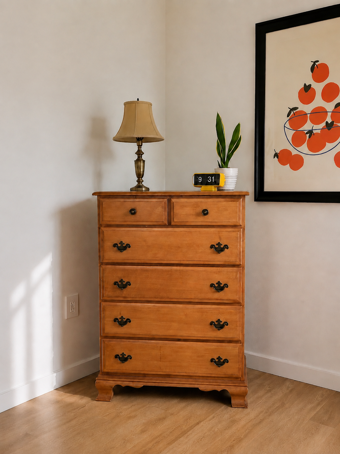 A wooden dresser with five drawers and decorative black handles stands against a white wall. On top, there is a brass table lamp with a beige shade, a potted plant with long green leaves, and a yellow flip clock displaying 9:31. To the right, a framed print of an abstract orange fruit design is hung on the wall.