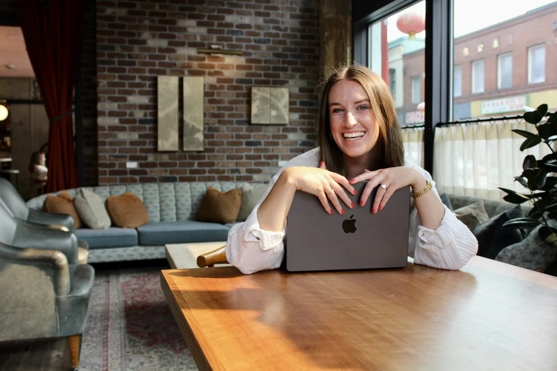 A woman sitting at a wooden table with a laptop, smiling, in a cozy cafe with a brick wall and large window behind her.