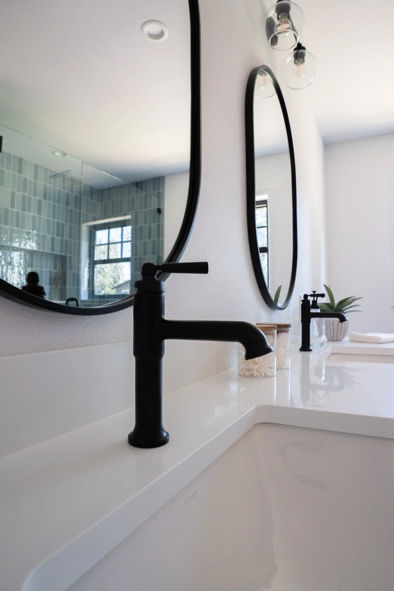 Close-up of a modern bathroom sink with a black faucet, round mirror, and transparent jars with cotton and soap, reflected in a large mirror. The bathroom has a minimalist design with white walls and a window with natural light.