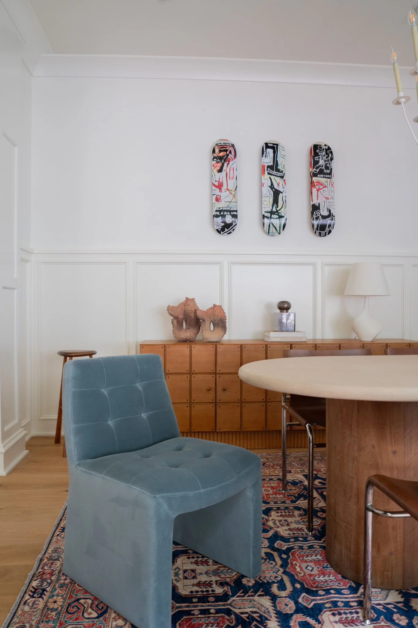 Living room with a blue upholstered chair, a wooden sideboard, decorative vases, a patterned rug, and three skateboards with graffiti art hanging on the white wall.