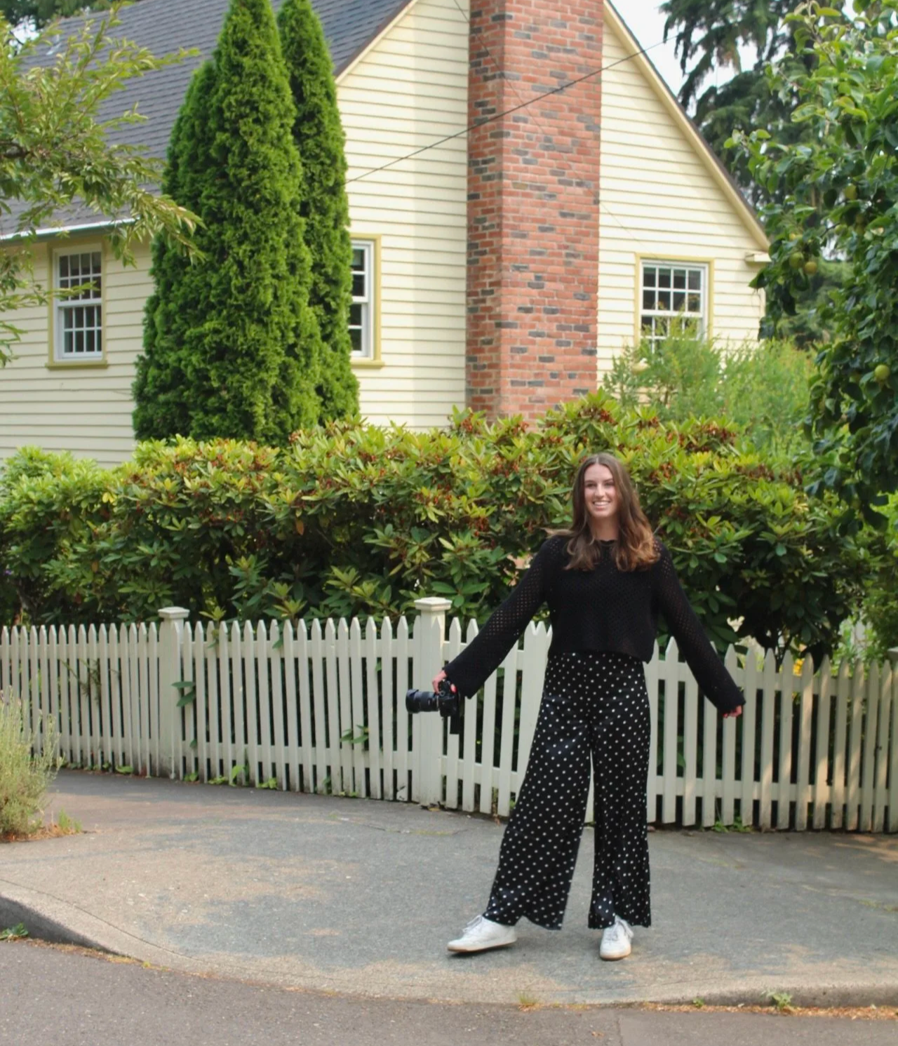 A young woman with long brown hair smiling and holding a curling iron in her right hand stands on a sidewalk in front of a white picket fence, green bushes, tall trees, and a beige house with a brick chimney.