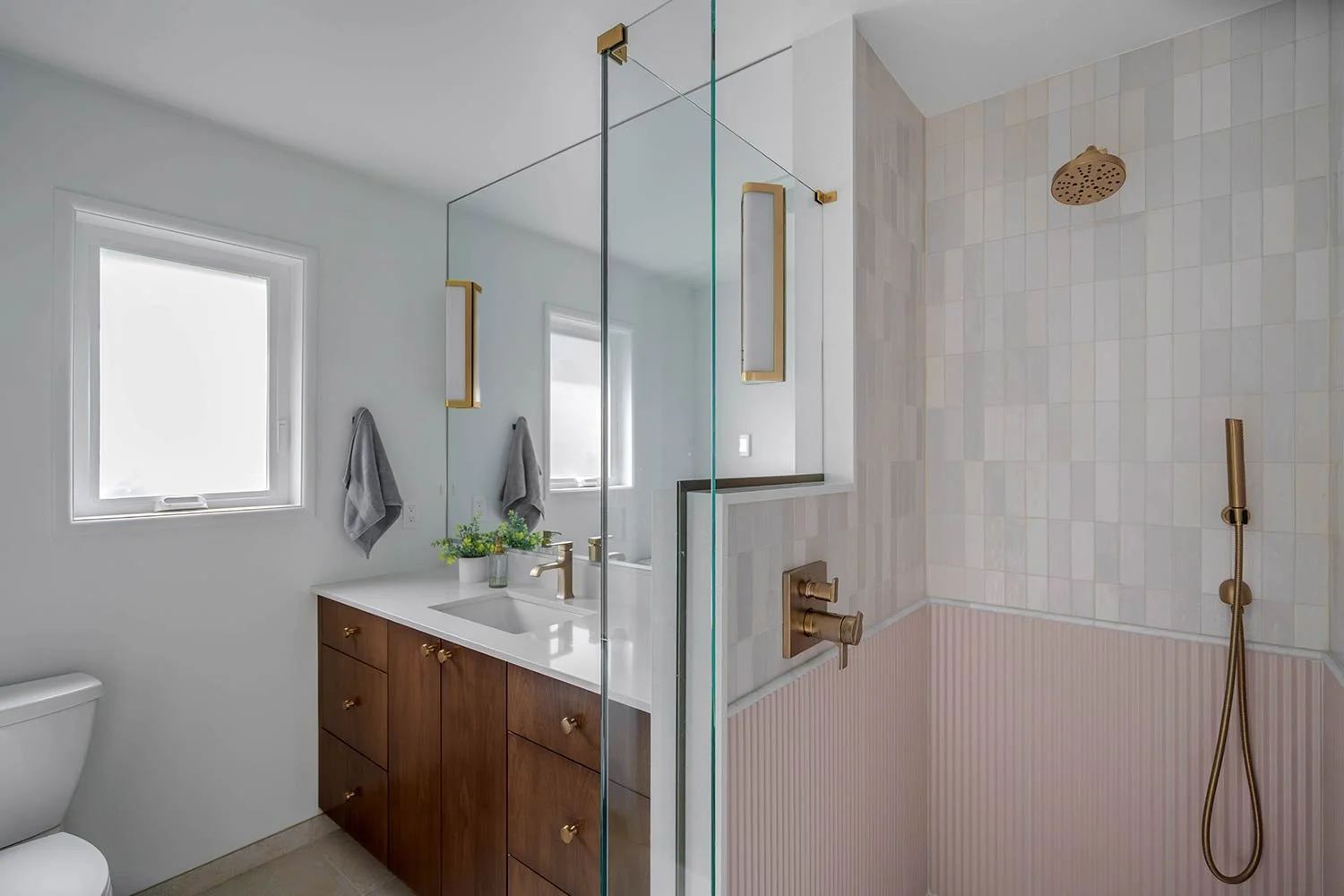 Modern bathroom with white walls, two small windows, a wooden vanity with a white countertop, a sink, a gold faucet, gray towels hanging on hooks, a potted plant, and a glass-enclosed shower area with beige and pink tiles, a gold showerhead, and a gold hand shower.