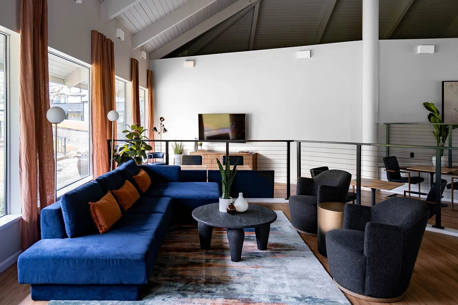 Modern living room with large navy blue sectional sofa, orange accent pillows, black armchairs, a round coffee table with potted plants and vases, wooden flooring, colorful rug, large windows with rust-colored curtains, and a wall-mounted television, with a dining area in the background.