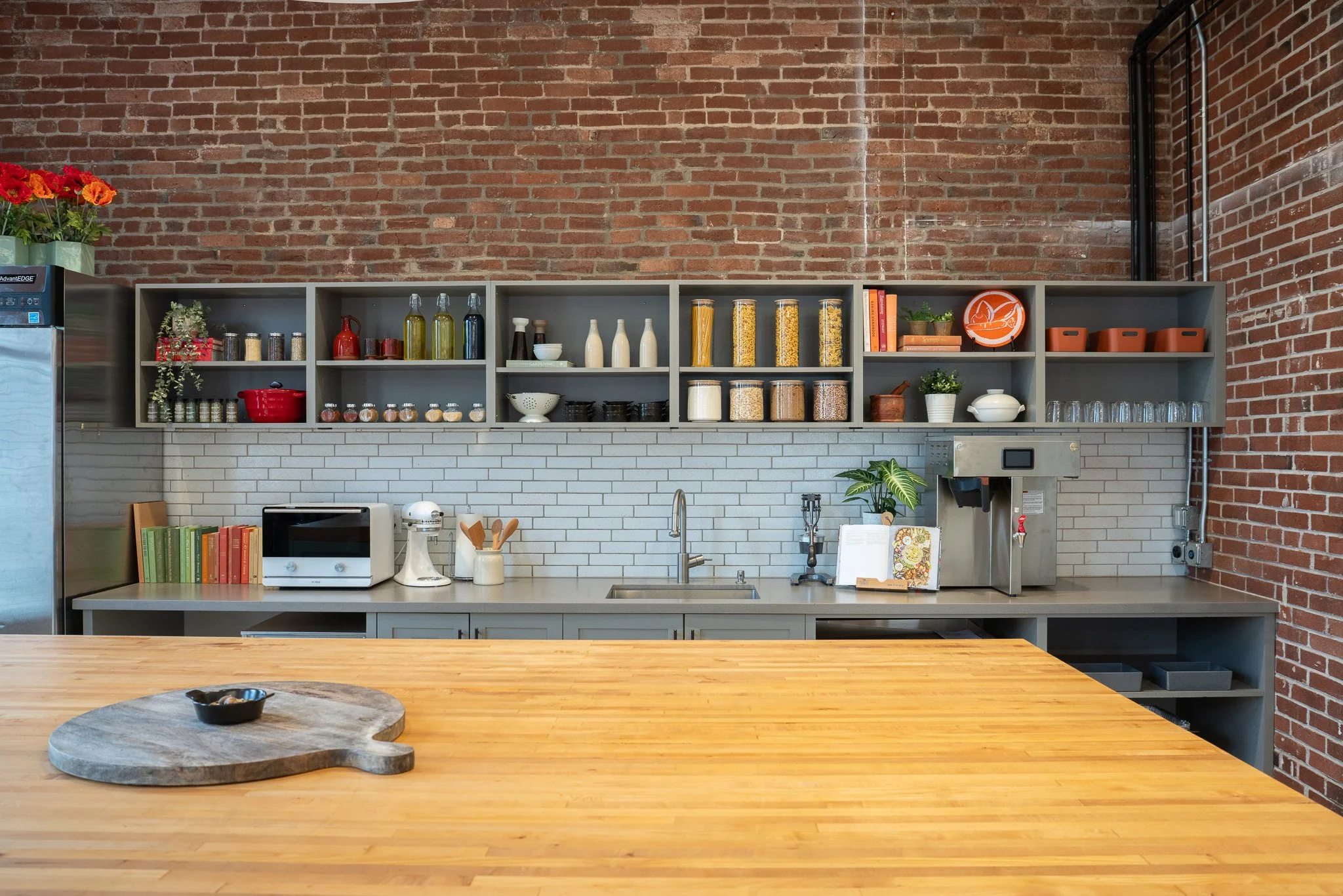 Kitchen with open shelving containing jars, books, plants, plates, and bottles against a brick wall, with a wooden table in the foreground.