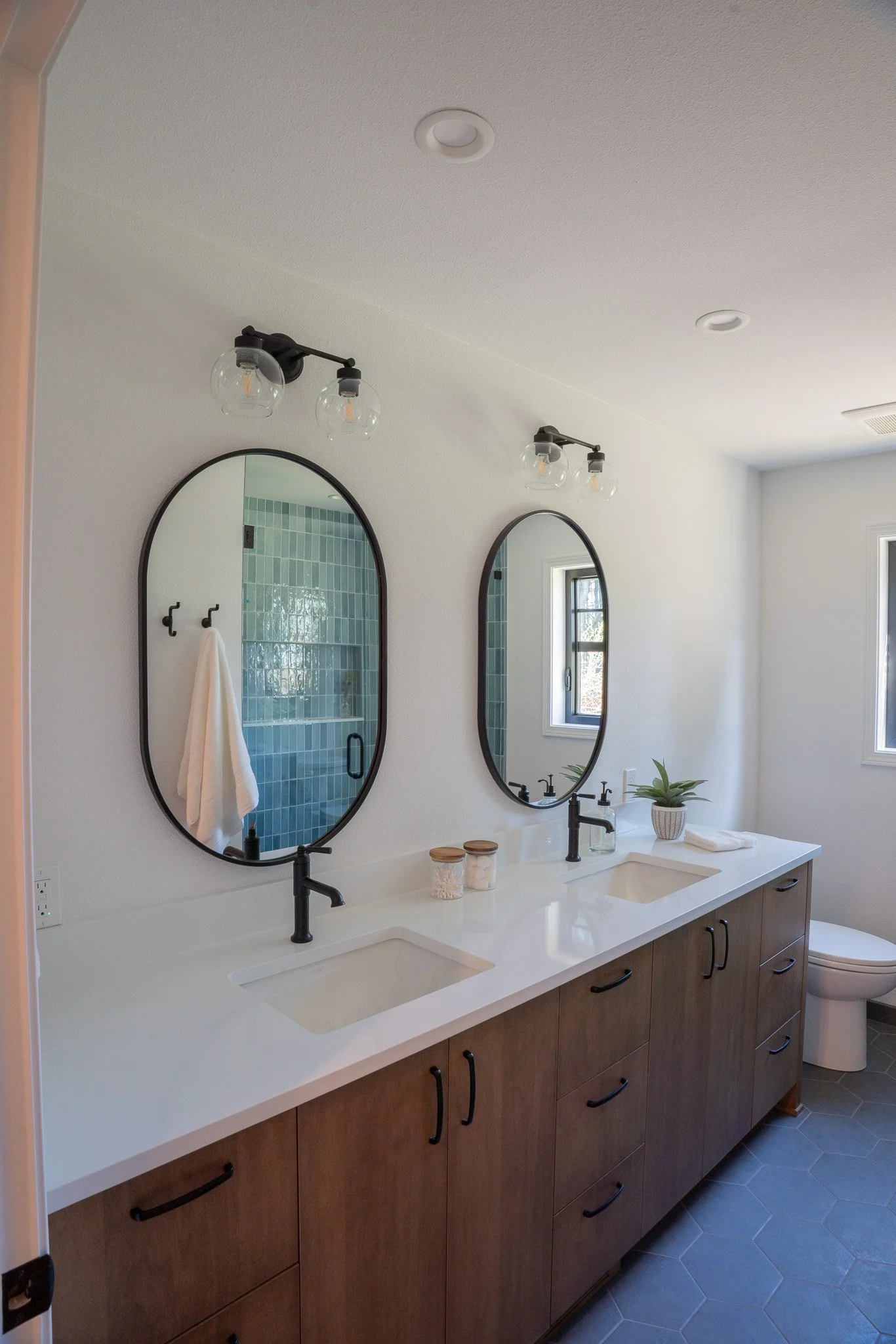 Bathroom with two large oval mirrors above a white vanity with dual sinks and black faucets, wooden cabinet drawers, and a potted plant. There are wall-mounted light fixtures with exposed bulbs, a window, and shower with blue tiles reflected in the m