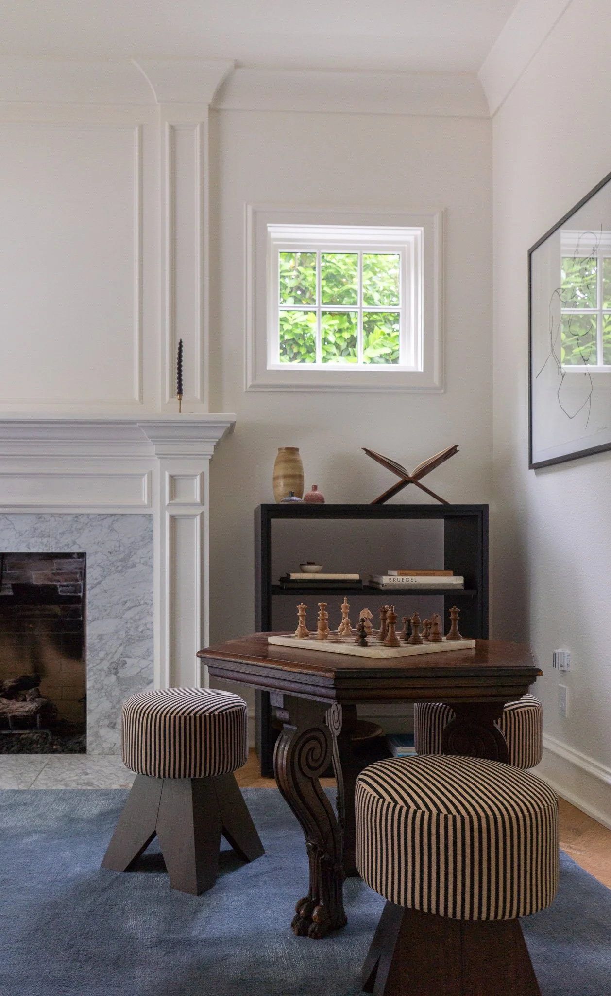 Interior of living room with a fireplace, a window with greenery outside, a small black bookshelf with decorative items, and a wooden table with chessboard and pieces. Two striped upholstered stools are visible, and a large framed abstract line drawi