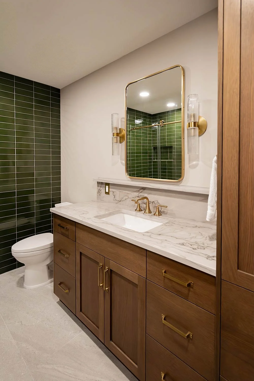 A modern bathroom with a wooden vanity, marble countertop, and gold fixtures. Green tiled wall and a large mirror above the sink. Toilet in the corner, and a shower area with a glass door reflected in the mirror.
