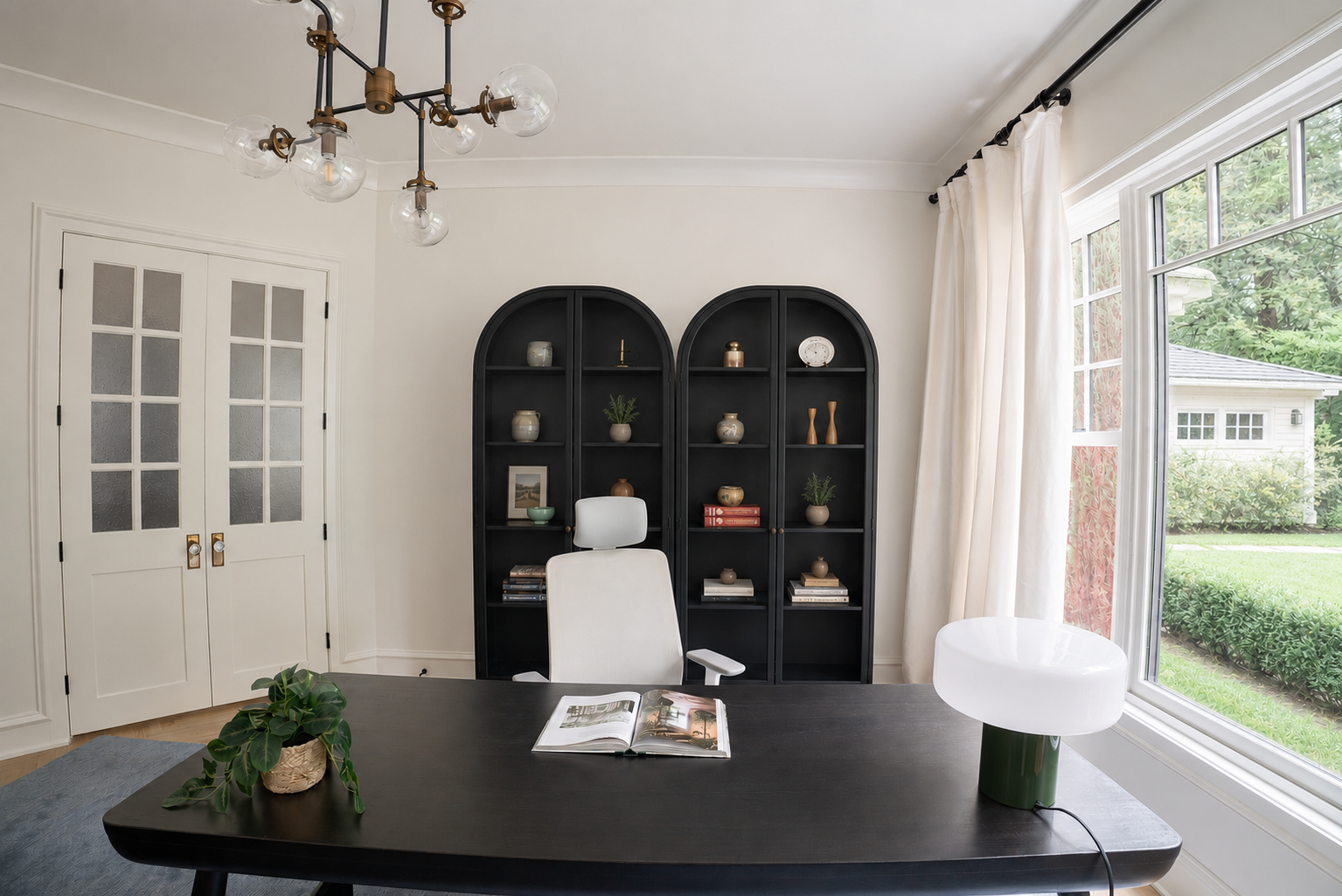 Home office with black bookshelf, white desk chair, dark desk, green plant, and a table lamp near large window with white curtains.