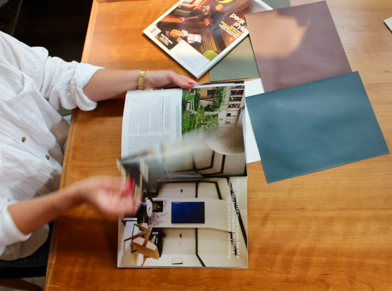 Person holding an open magazine showing interior design images, with pages displaying a modern living room, on a wooden table surrounded by other magazines and colored paper sheets.