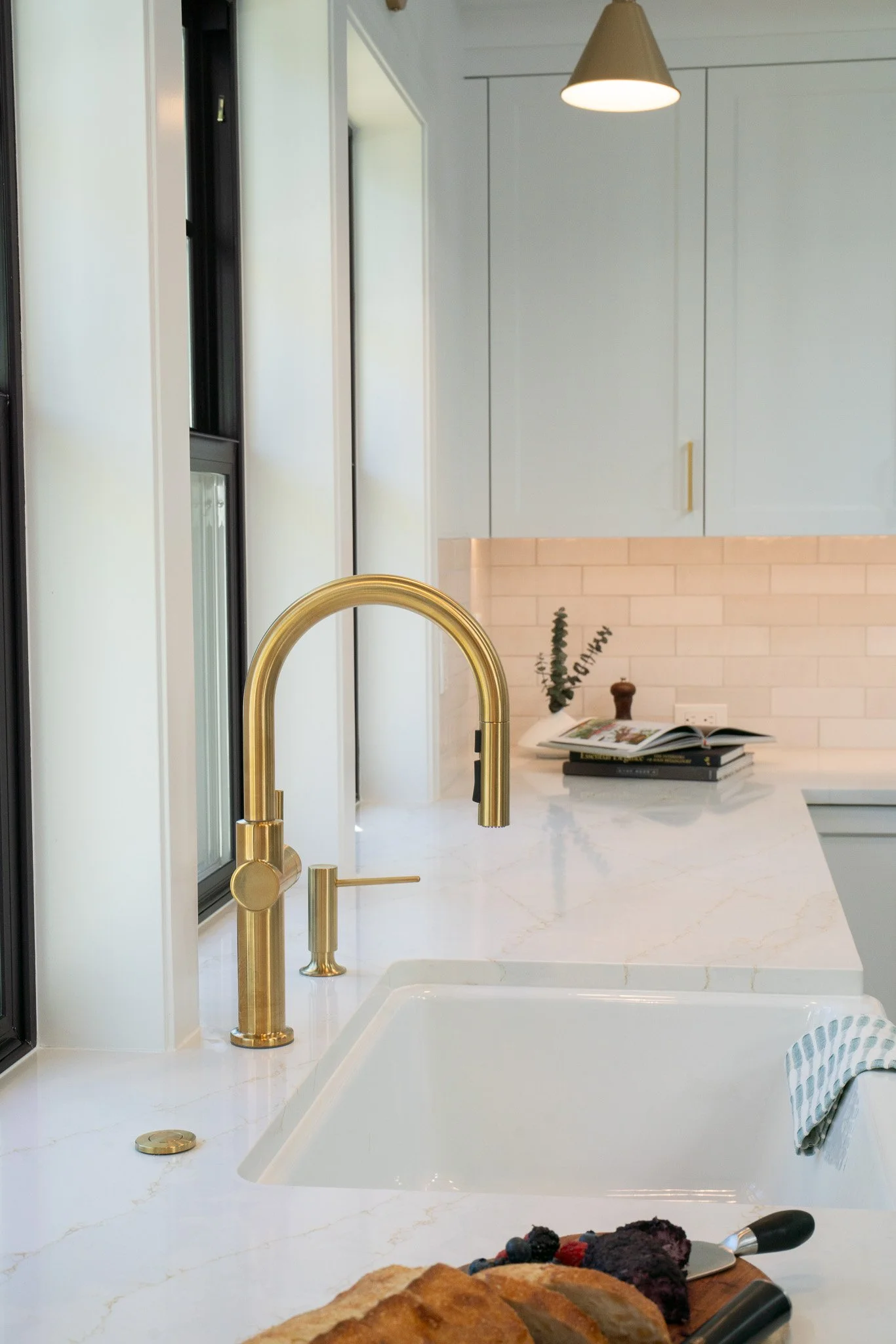 A kitchen countertop with a white marble surface and a built-in sink. A gold faucet is installed, with a hand towel hanging on the right side. To the side, there is a cutting board with baked bread and berries, and a bread knife. In the background, t