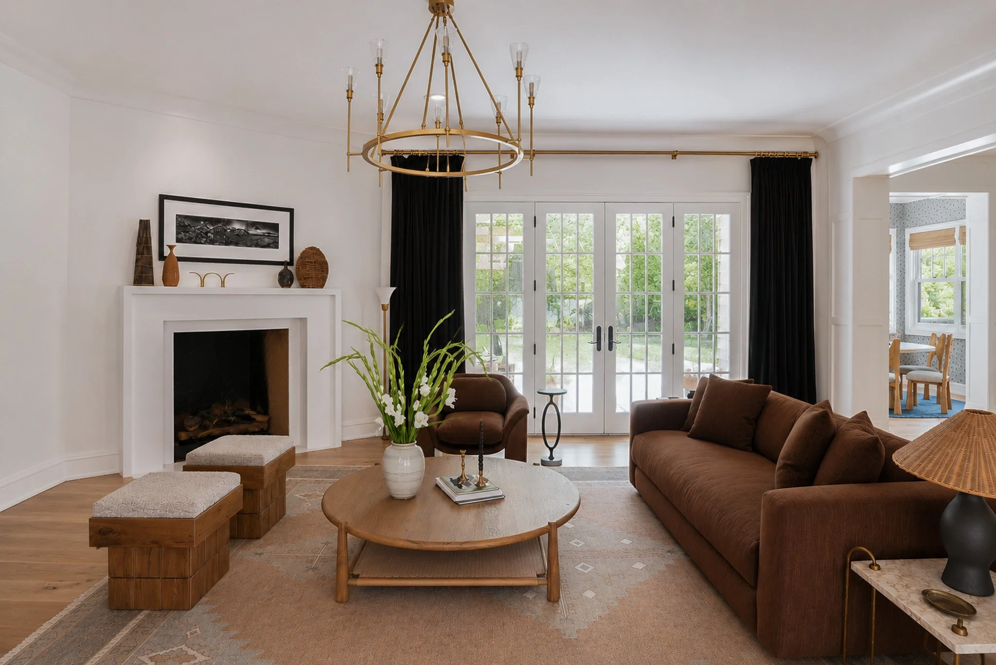 Living room with white walls, a fireplace, wooden flooring, and large windows. A brown sofa, a round wooden coffee table, and potted plants are present, with decorative items on the mantle and side tables. Black curtains frame the windows, and there'