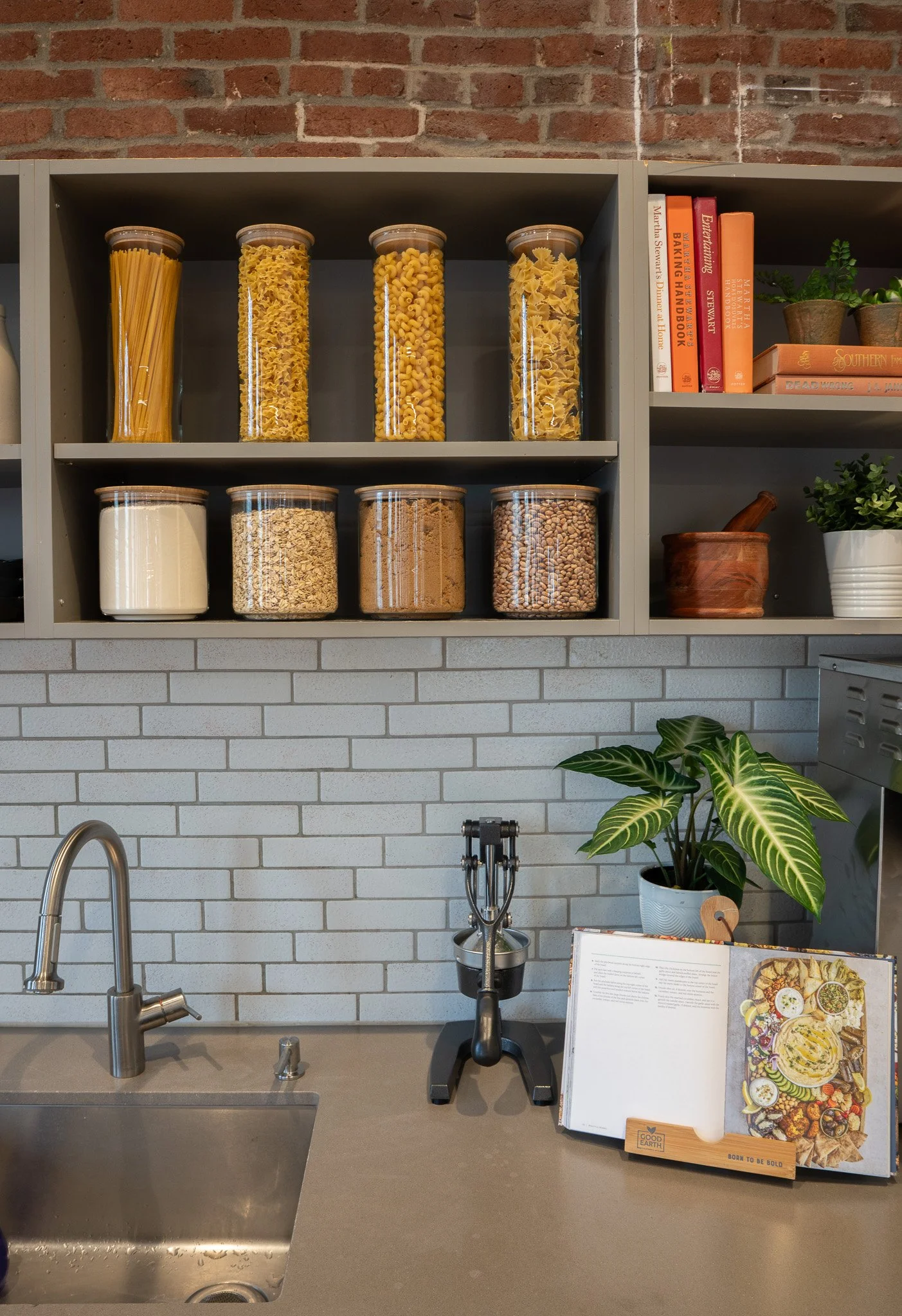 Kitchen with a modern gray countertop, a stainless steel sink, a black faucet, open shelves with glass containers of pasta and grains, books about baking and entrepreneurship, potted plants, and a cookbook on a wooden stand.