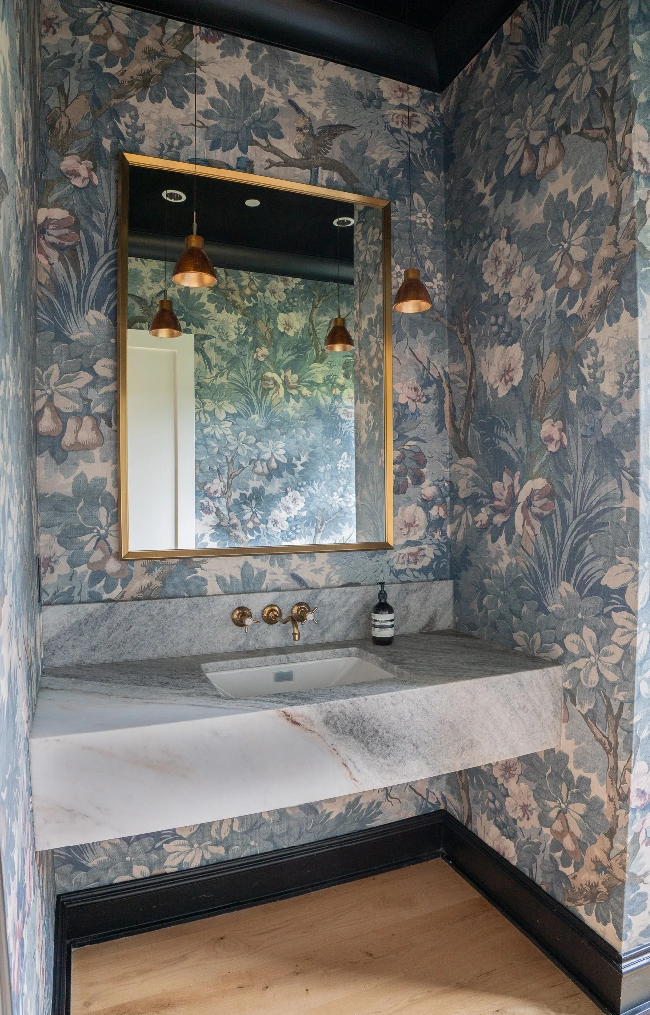 A bathroom vanity with a rectangular mirror, floral-patterned wallpaper, a marble countertop with an undermount sink, gold fixtures, and three hanging copper pendant lights.
