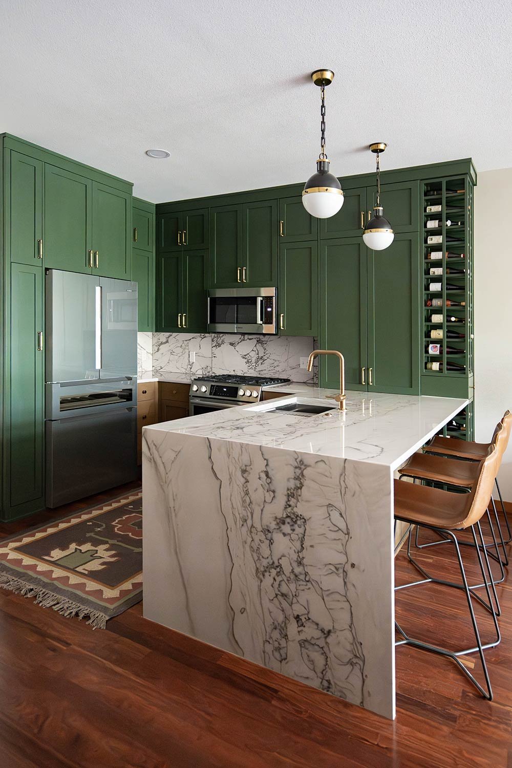 Modern kitchen with green cabinets, marble island, stainless steel appliances, wine storage, pendant lighting, and leather barstools.
