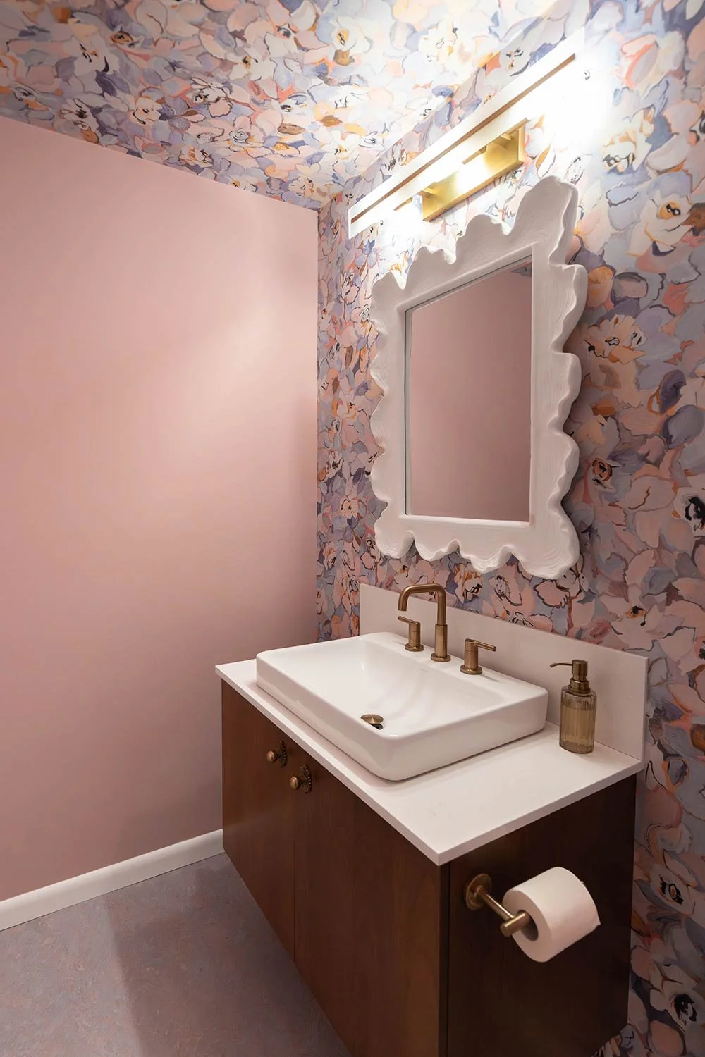 A bathroom with a pink wall on the left and a floral patterned wallpaper on the right, featuring a large scalloped white-framed mirror, a white vessel sink with gold fixtures, a small soap dispenser, and a gold toilet paper holder with a roll of toilet paper.