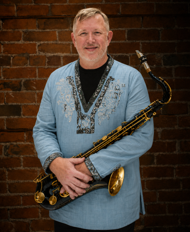 A middle-aged man with short blond hair and a beard holding a black and gold saxophone, standing in front of a red brick wall, wearing a light blue embroidered shirt.