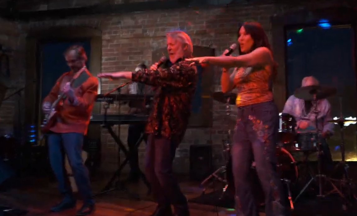 A band performing on stage with three women singing and playing instruments, in a venue with brick walls and colorful lighting.