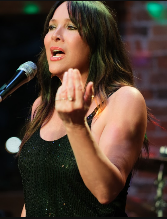A woman with long brown hair singing into a microphone on stage, wearing a black, sparkly sleeveless top, with a brick wall background.