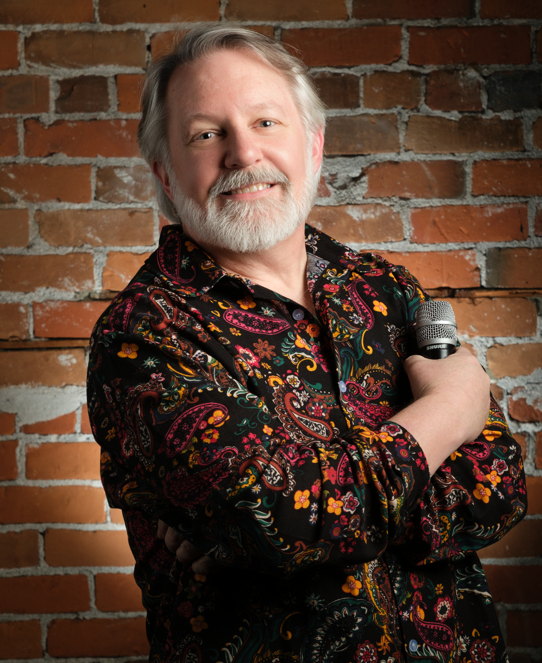 A man with gray hair and beard holding a microphone, smiling, standing in front of a brick wall, wearing a colorful paisley shirt.