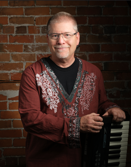 A middle-aged man with glasses smiling, standing in front of a brick wall, wearing a maroon shirt with floral embroidery, holding a keyboard.