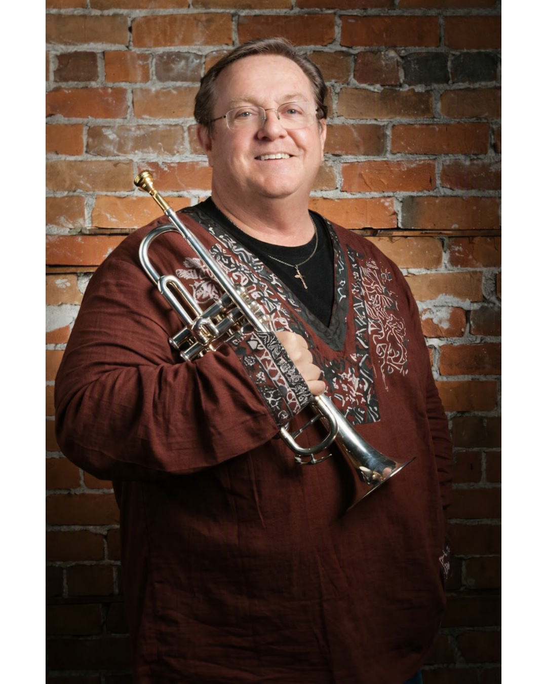 A smiling man standing in front of a brick wall, holding a trumpet over his shoulder. He is wearing glasses, a black shirt, a maroon shirt with embroidery, and a necklace with a cross.