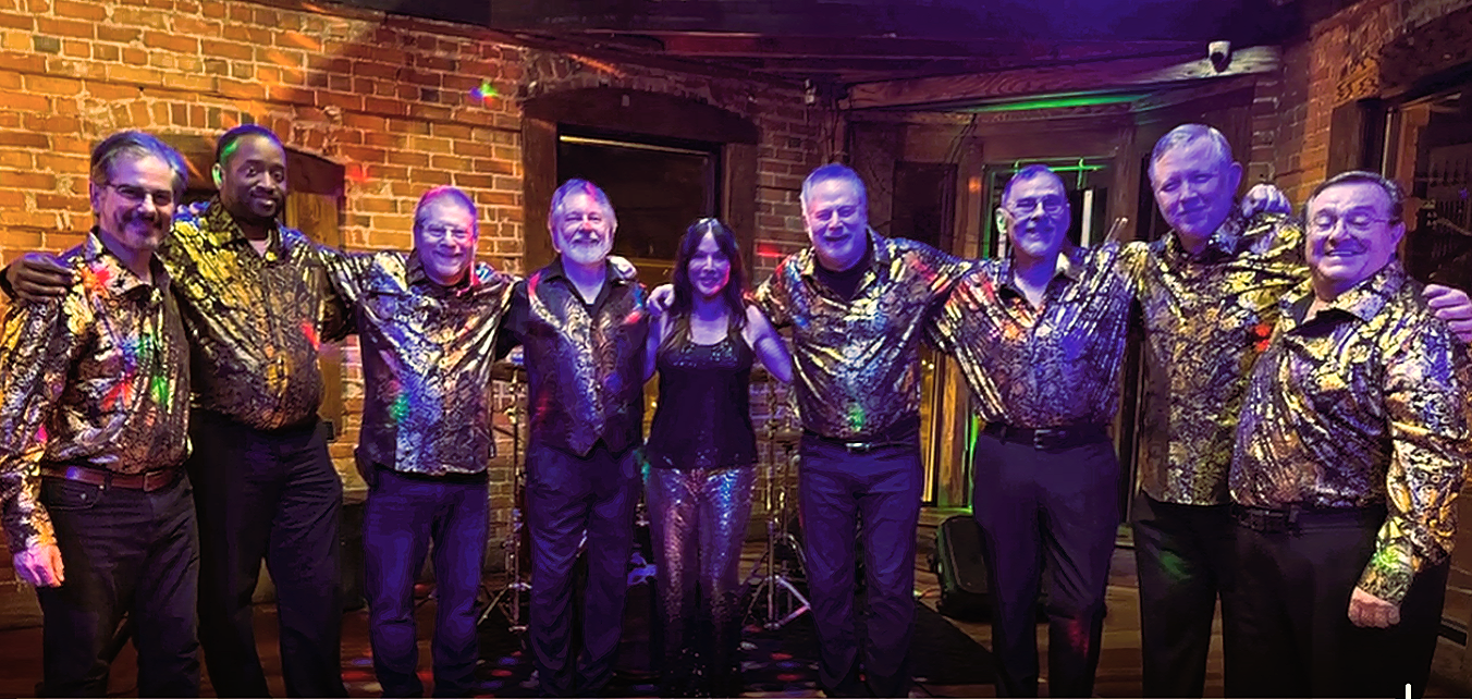 A group of ten people, nine men and one woman, stand together with arms around each other in a brick-walled room, dressed in matching shiny, colorful, patterned shirts. A woman in the center wears a black top and shiny pants. The setting appears to be a lively indoor event or party with musical instruments visible in the background.