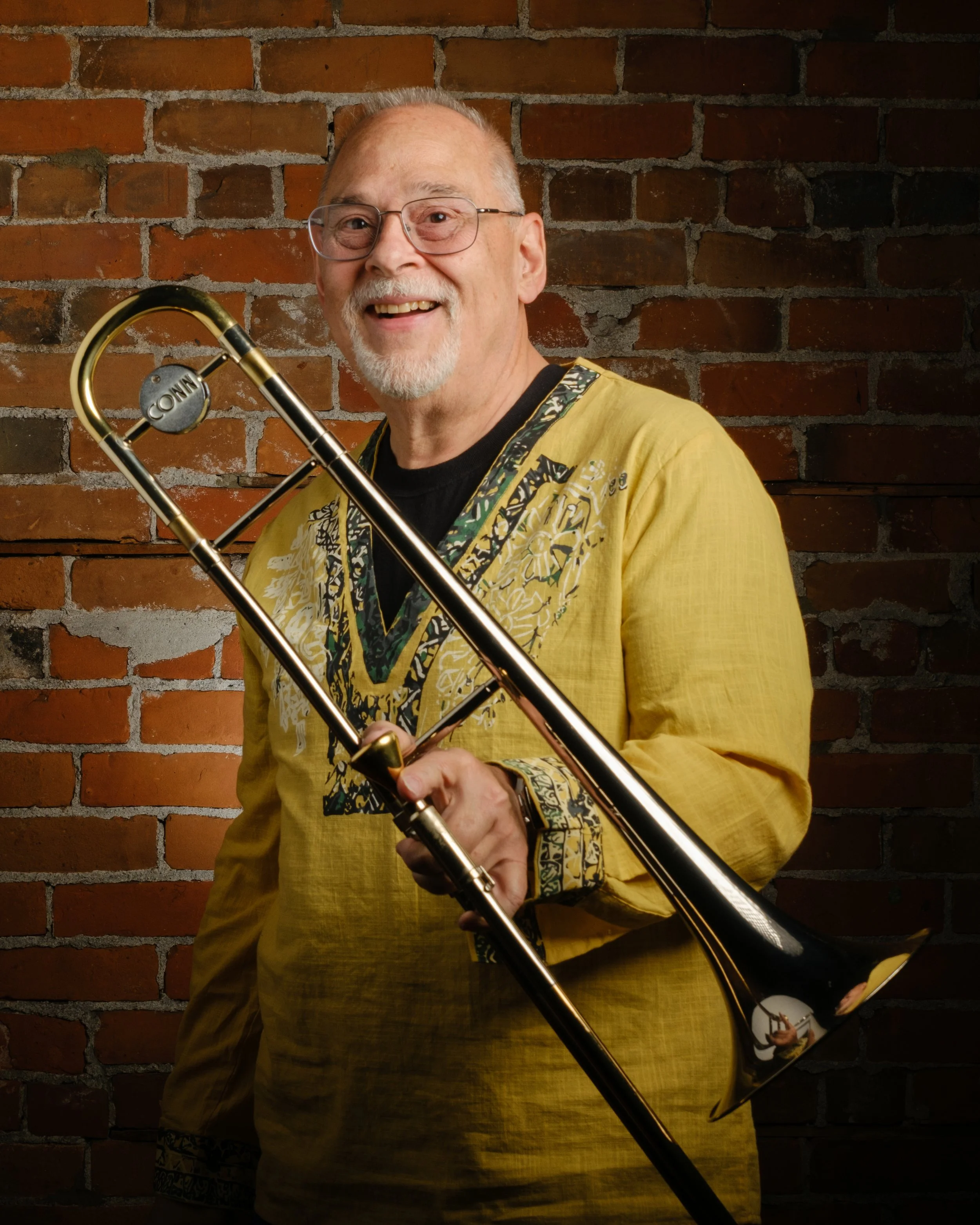 An elderly man with glasses, a mustache, and a beard is smiling while holding a trombone. He is wearing a yellow shirt with a decorative black and gold pattern on the collar and sleeves. The background is a brick wall.