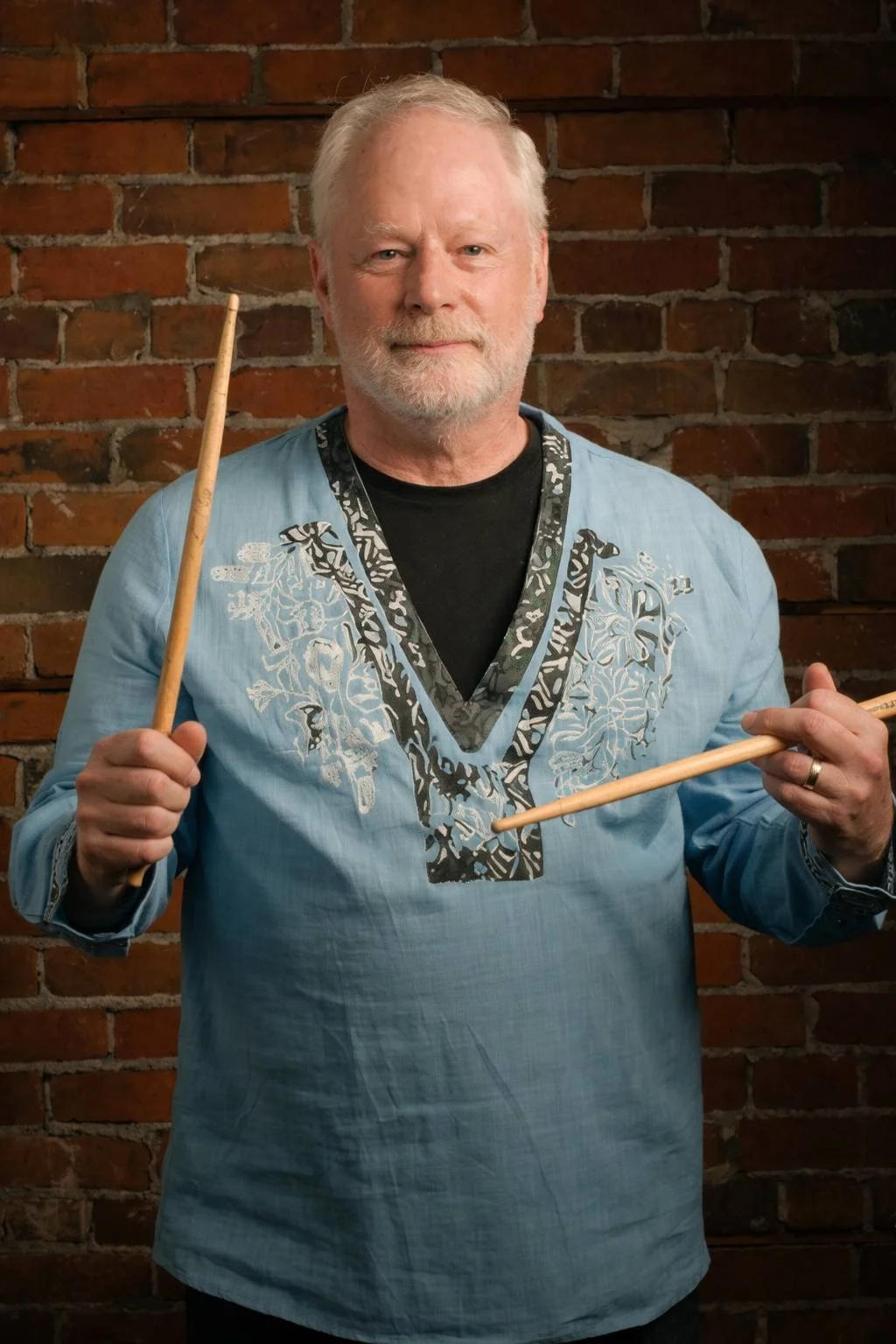 A man with gray hair and beard holding a drumstick in each hand, standing in front of a brick wall, wearing a blue embroidered shirt over a black t-shirt.