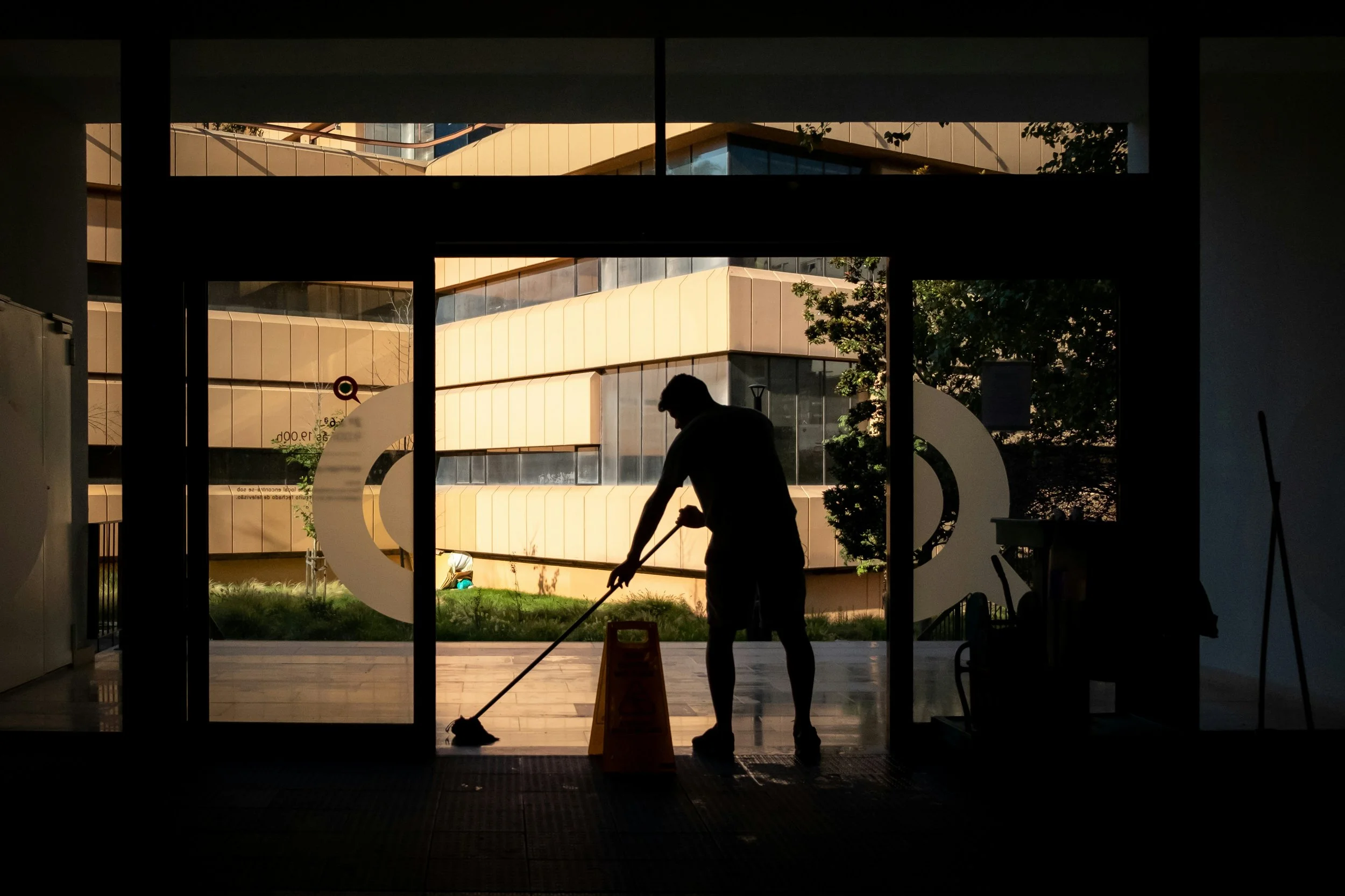 Une personne en silhouette nettoie le sol à l'intérieur d'un bâtiment très propre, vue à travers une grande porte vitrée propre, avec un bâtiment moderne en verre et acier visible à l'extérieur.