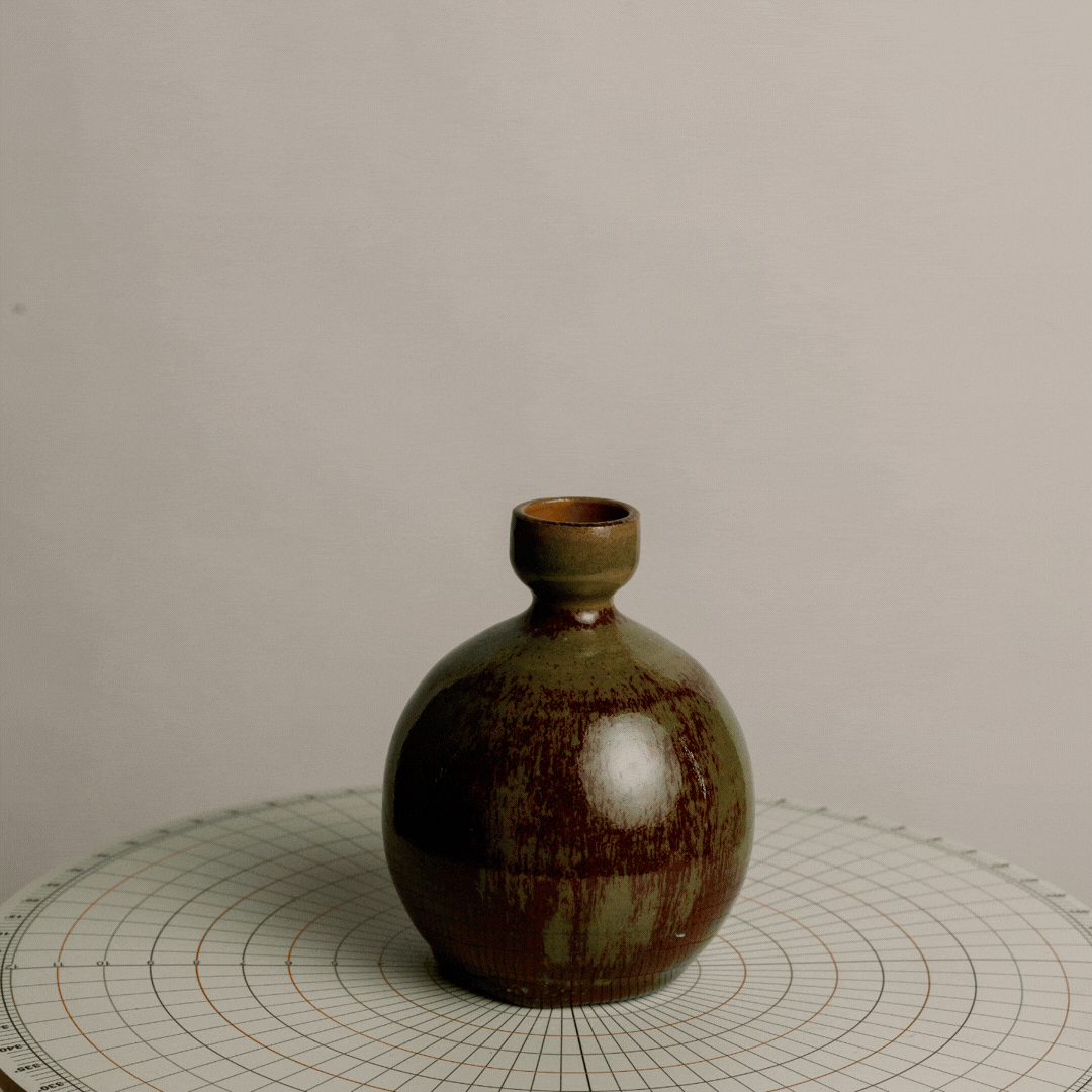 A small, round ceramic vase with a narrow opening, placed on a round grid-patterned surface against a plain background.
