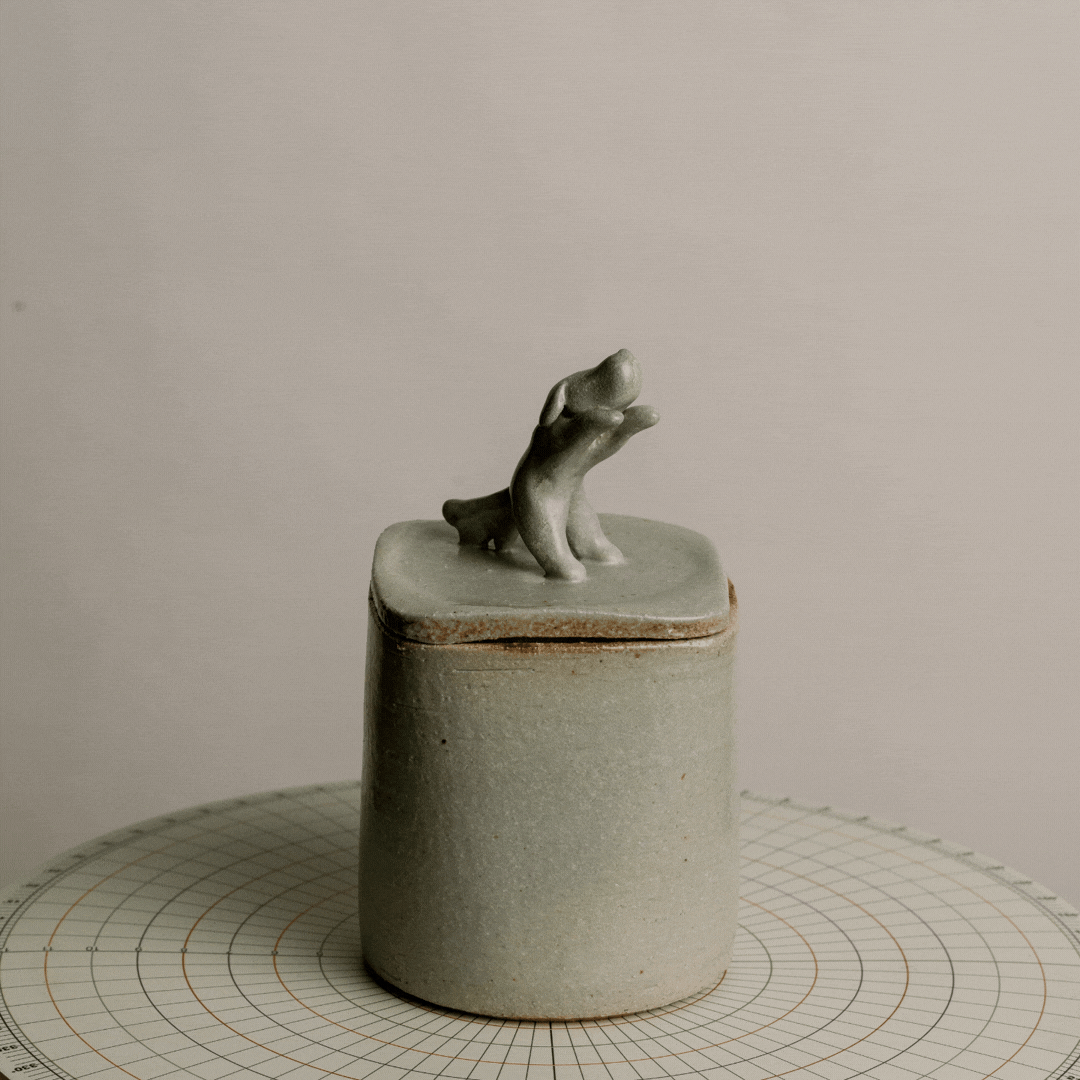 A small, gray ceramic container with a lid featuring a tiny sculpture of a dog on top, placed on a round, grid-patterned surface.