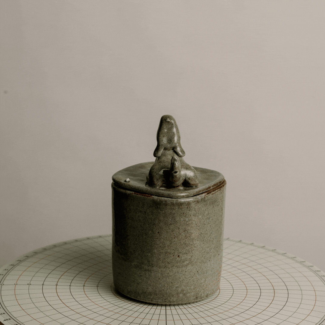 A ceramic container with a lid featuring a rabbit figurine on top, placed on a grid-patterned surface.