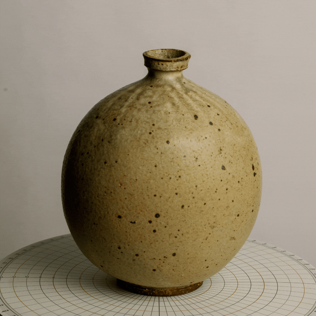 A round beige ceramic vase with small dark speckles, narrow opening at top, placed on a circular grid-patterned surface.