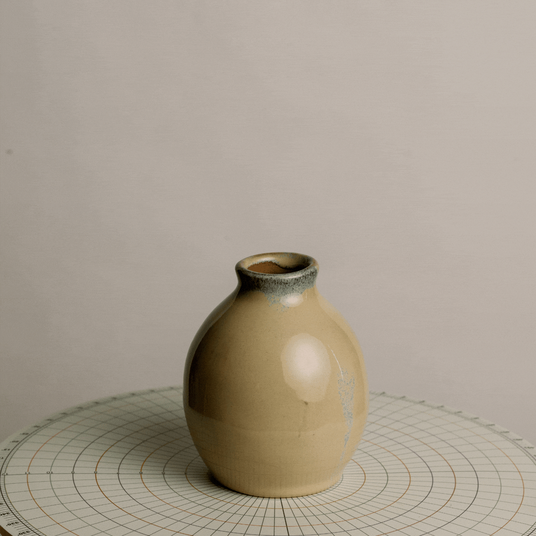A beige ceramic vase with a slightly flared opening, placed on a circular grid-patterned surface against a plain background.