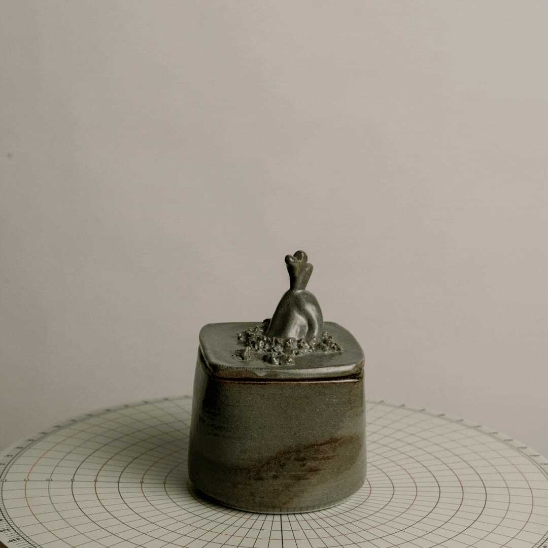 A ceramic container with a sculpture of a rabbit on top, placed on a grid-lined surface.