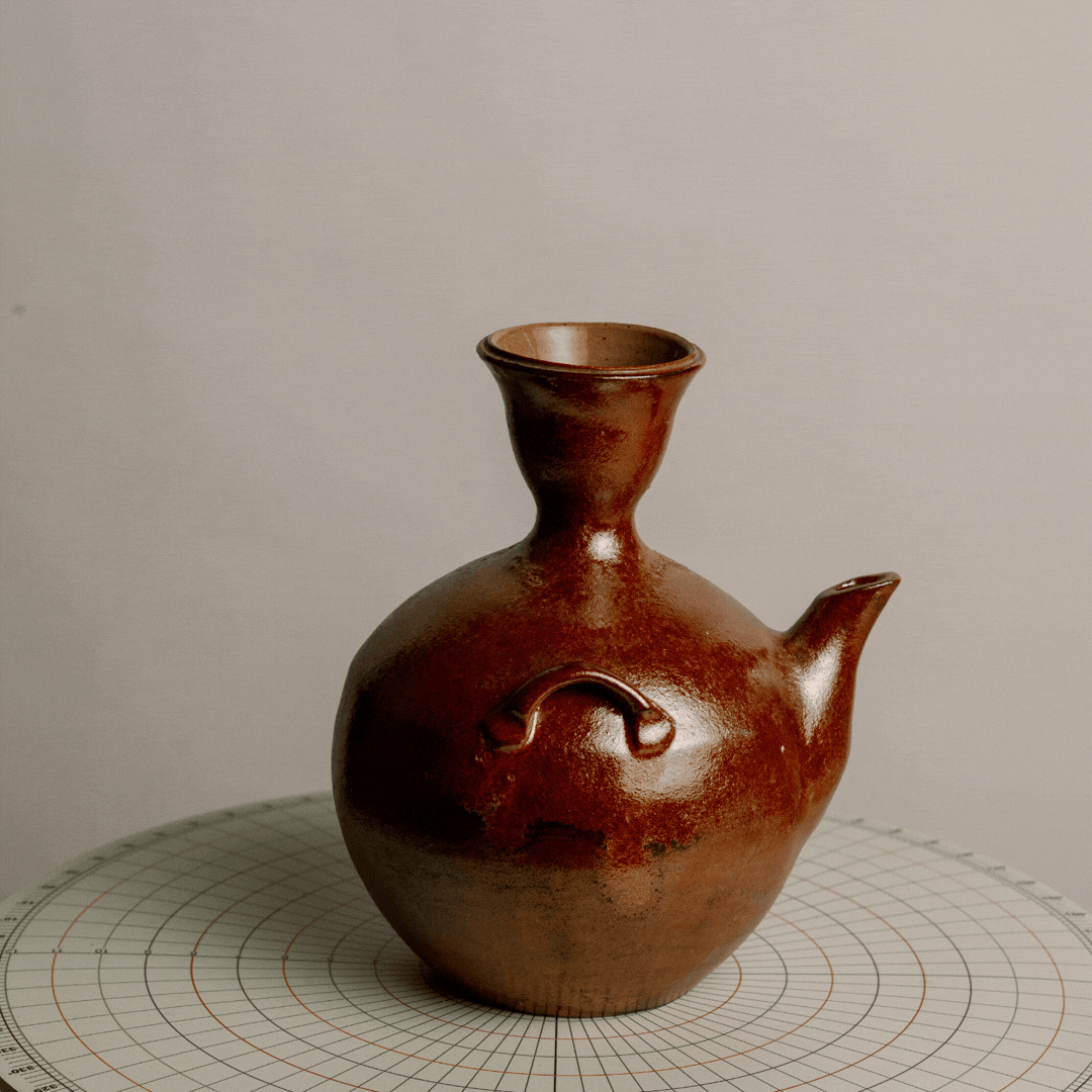 A brown ceramic vessel shaped in the form of a teapot with a narrow neck, spout, and decorative handle, placed on a round patterned surface.