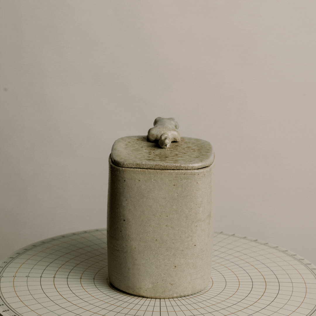 A cylindrical ceramic container with a lid, featuring a small, decorative animal figure on top, placed on a round, gridded surface.