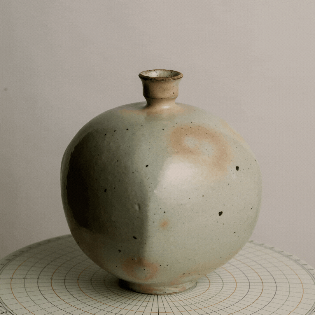 A round ceramic Vase with a small opening at the top, featuring a minimalist and textured design in pale green and beige tones, placed on a grid-patterned surface.