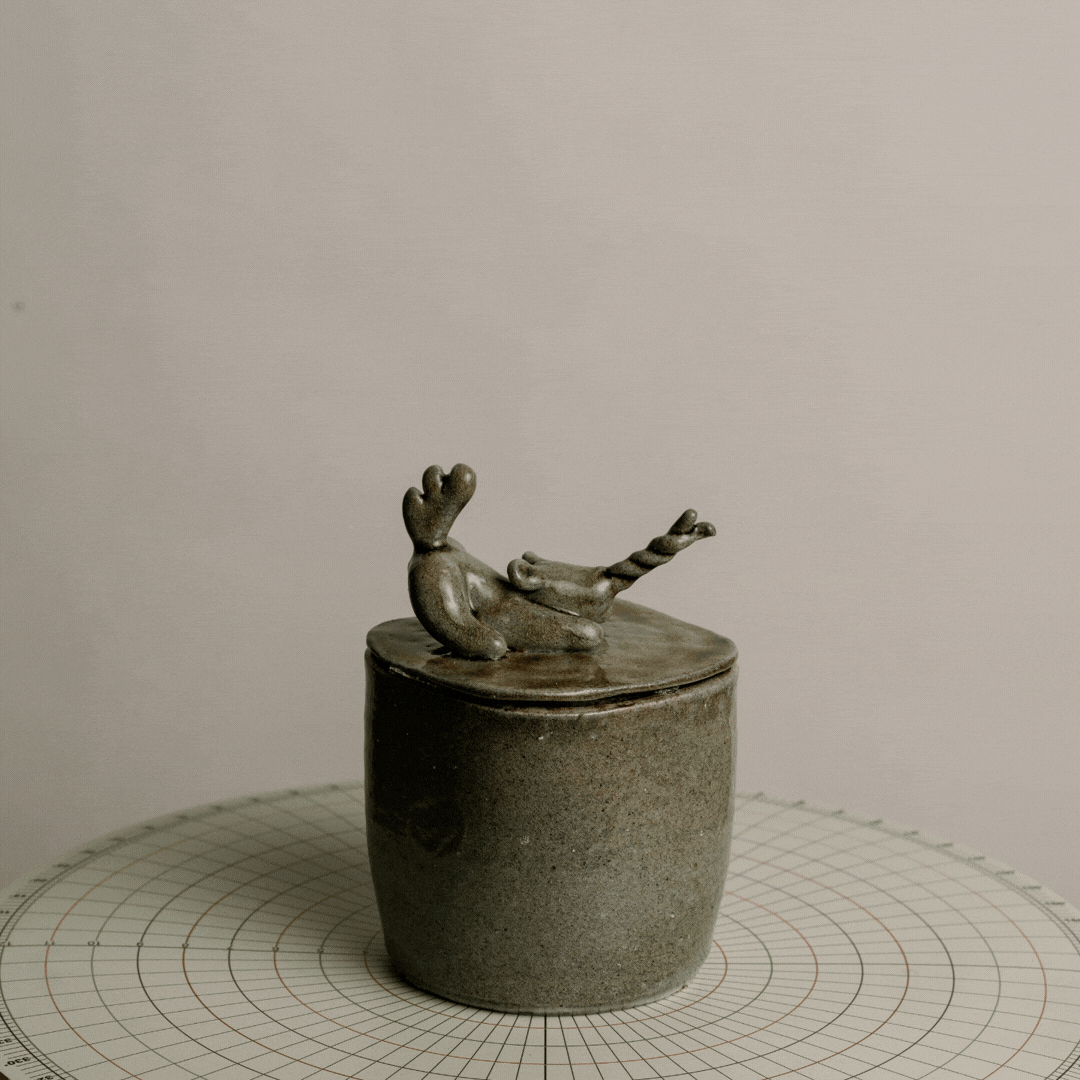 A small gray ceramic container with a lid, featuring a decorative ceramic deer sitting on top of the lid.