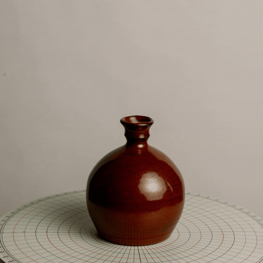 A small brown ceramic vase with a narrow neck, placed on a round, grid-patterned surface against a plain gray background.