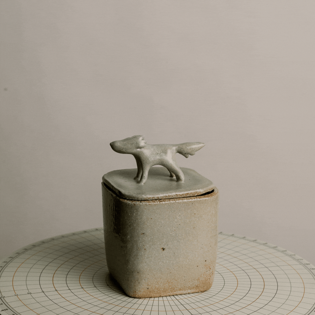A small ceramic container with a lid, topped by a decorative dog figurine, placed on a round, grid-lined surface.