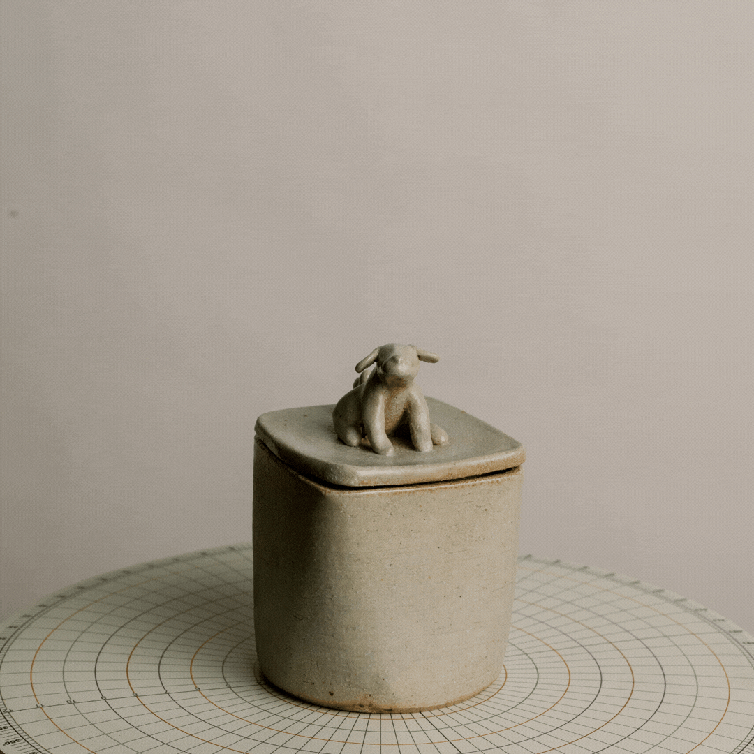 A small ceramic container with a lid, decorated with a tiny rabbit figurine on top, placed on a white grid-patterned surface.